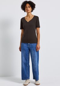 Tmavě hnědé tričko s výstřihem do V a krátkými rukávy, v kombinaci s světle modrými širokými džíny; model obutý v béžových teniskách, stojící na jednobarevném pozadí.
