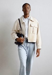 Giacca cropped beige con due grandi tasche frontali, indossata sopra una camicia a righe azzurra chiara, abbinata a jeans denim a vita alta.