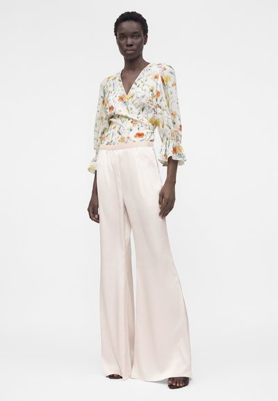 Γυναίκα που φοράει λευκό σατέν παντελόνι με φαρδιά πόδια και λευκή floral τοπ τύπου περιτύλιξης με 3/4 μανίκια, στέκεται μπροστά σε ένα απλό λευκό φόντο.