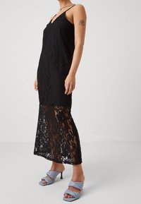 Black lace dress with a V-neck and spaghetti straps, featuring a sheer lace pattern at the bottom. Paired with blue heeled sandals.