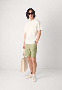 Cremefarvet t-shirt med struktur og brystlomme, grønne shorts og en lys tote bag, båret med hvide sneakers og sorte solbriller.