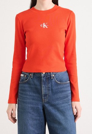 Femme portant un haut orange Calvin Klein ajusté à manches longues et un jean bleu taille haute, debout les bras détendus.