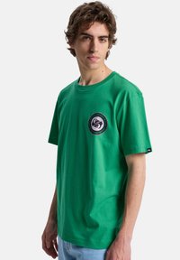 Jeune homme aux cheveux bruns ondulés portant un T-shirt vert avec un logo circulaire sur la poitrine et un jean bleu clair, debout sur un fond blanc.