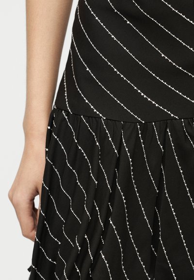 Μαύρο φόρεμα με ασημένιες διαγώνιες ρίγες από παγιέτες, με εφαρμοστή μέση και πλισέ τμήμα φούστας, χέρι ακουμπισμένο δίπλα στο φόρεμα.