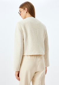 Veste crème cropped en tissu texturé et duveteux, avec de longues manches et un col rond simple. Associée à un pantalon beige clair.