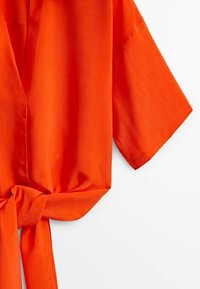 Blouse courte à manches courtes de couleur orange vif avec un col en V et un nœud noué à l'avant, présentée sur un fond blanc.