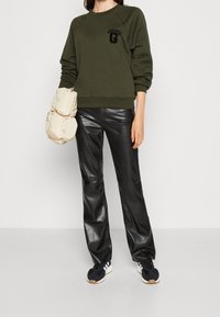 Persona che indossa una felpa verde oliva con logo, pantaloni in ecopelle nera, sneakers bianche e nere, mentre tiene una borsa imbottita beige.