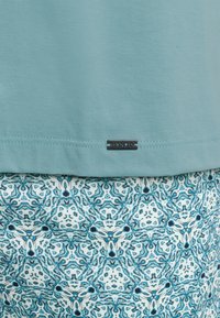 Top in cotone azzurro chiaro con una piccola etichetta nera; abbinato a un tessuto stampato con motivi geometricali blu e bianchi.