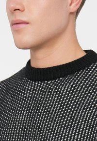 Maglione lavorato a maglia in bianco e nero con un motivo testurizzato, caratterizzato da un colletto nero a coste. Inquadratura ravvicinata del collo e della spalla di un modello.