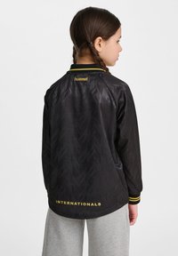 Svart jacka med ett subtilt geometriskt mönster, guld "hummel"-logotyp på kragen och texten "INTERNATIONALS" vid fållen. Har gula ränder vid ärmsluten.