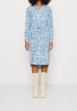 Kobieta ubrana w jasnoniebieską sukienkę koszulową z długim rękawem, ozdobioną czarnymi i beżowymi półksiężycowymi wzorami, z przewiązanym w talii paskiem, oraz kremowymi, sznurowanymi kozakami za kolano.