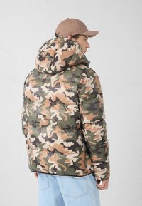 Giacca imbottita mimetica con cappuccio, caratterizzata da varie tonalità di verde, marrone e beige. Texture liscia e design trapuntato per il calore.