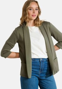 Blazer vert olive avec un design ajusté, devant ouvert et deux poches avant, associé à un t-shirt blanc et un jean bleu taille haute.