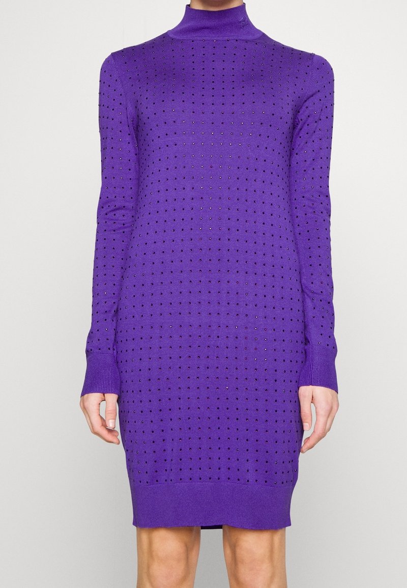 Robe en maille violette à manches longues avec de petites perforations régulièrement espacées et un col montant, portée par une personne debout.