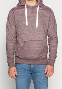 Hombre vestido con una sudadera con capucha color burdeos y cordones blancos, usando jeans azules, de pie frente a un fondo claro y simple.