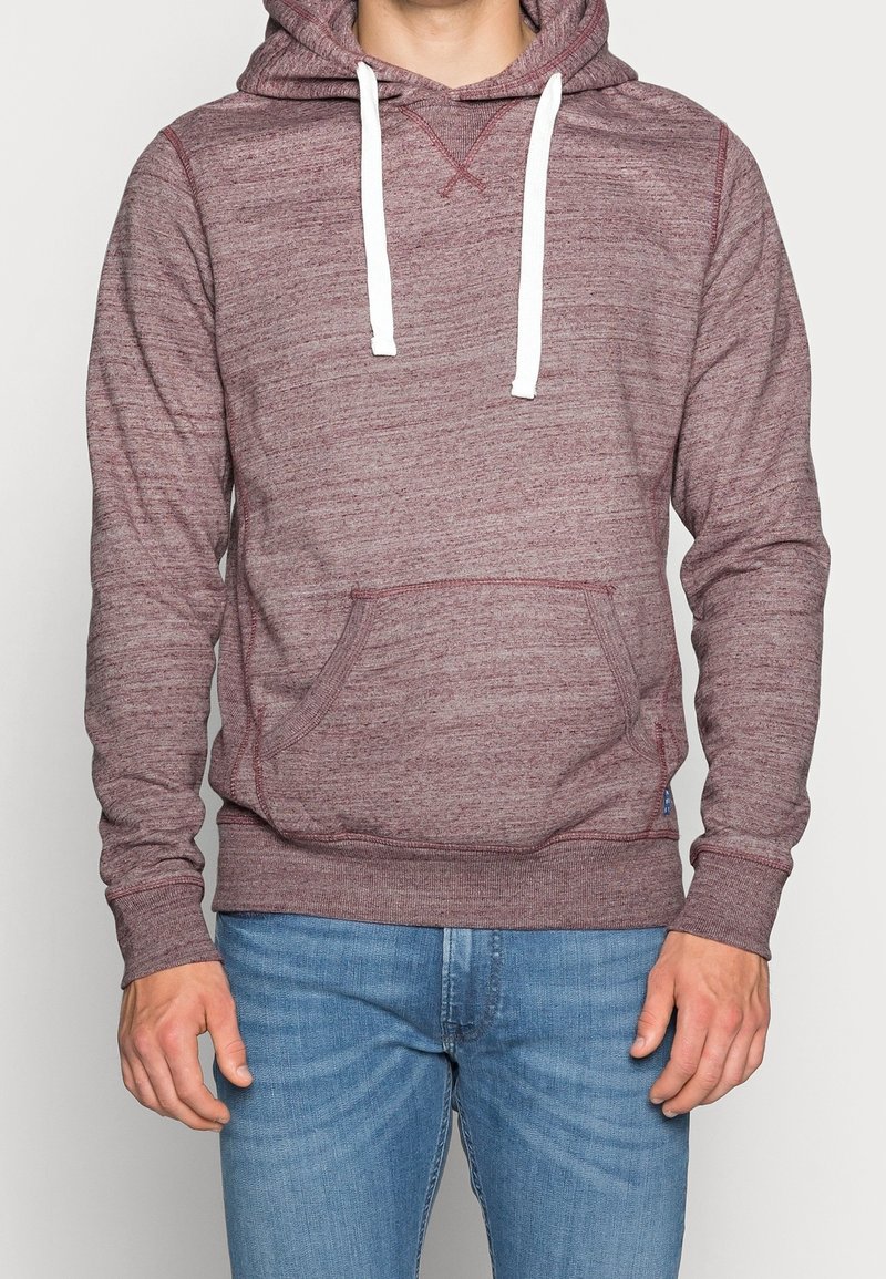 Hombre vestido con una sudadera con capucha color burdeos y cordones blancos, usando jeans azules, de pie frente a un fondo claro y simple.