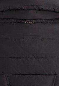 Svart vadderad jacka med kviltad textur, synlig sömnad och metall-logotyp som accent. Slät material med horisontella paneler.