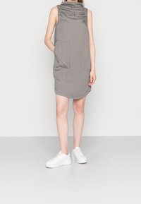 Vestido gris sin mangas con un alto cuello y dos bolsillos delanteros, hecho de una tela lisa. Combinado con zapatillas de plataforma blancas.