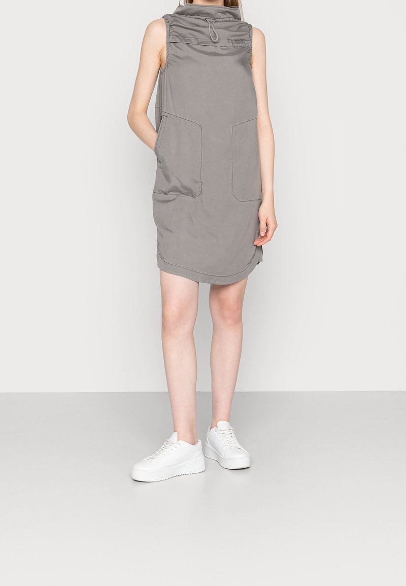 Vestido gris sin mangas con un alto cuello y dos bolsillos delanteros, hecho de una tela lisa. Combinado con zapatillas de plataforma blancas.