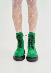 Botas de tobillo verde de ante con cordones, suelas de goma negra gruesa y detalles texturizados, combinadas con medias de red blancas con un patrón de lazo.