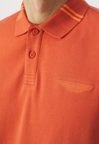 Oranssi polo-paita, jossa on teksturoitu kudos, kolme nappia ja kaulus, jossa on kaksi oranssia raitaa. Brodeerattu Aston Martin -logo rinnassa.