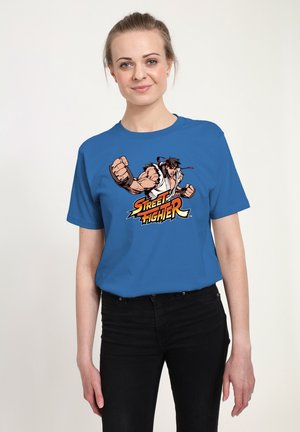 Kvinde iført en blå t-shirt med en muskuløs Street Fighter-figur og logo, kombineret med sorte bukser, stående foran en enkel hvid baggrund.