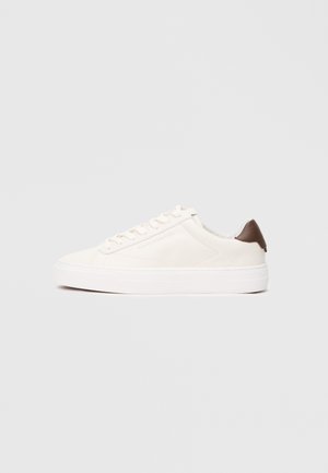 Sneaker bianca con design a lacci e linguetta marrone sul tallone, su uno sfondo chiaro uniforme.