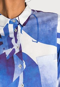 Kraťasová košile s modro-fialovo-bílým abstraktním vzorem, zdobená bílými knoflíky a kapsou na prsou na pravé straně.