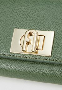 Grüne Lederhandtasche mit einem auffälligen goldenen Verschluss mit dem Buchstaben "A", strukturiertem Oberflächenmaterial und sichtbaren Nähdetails.