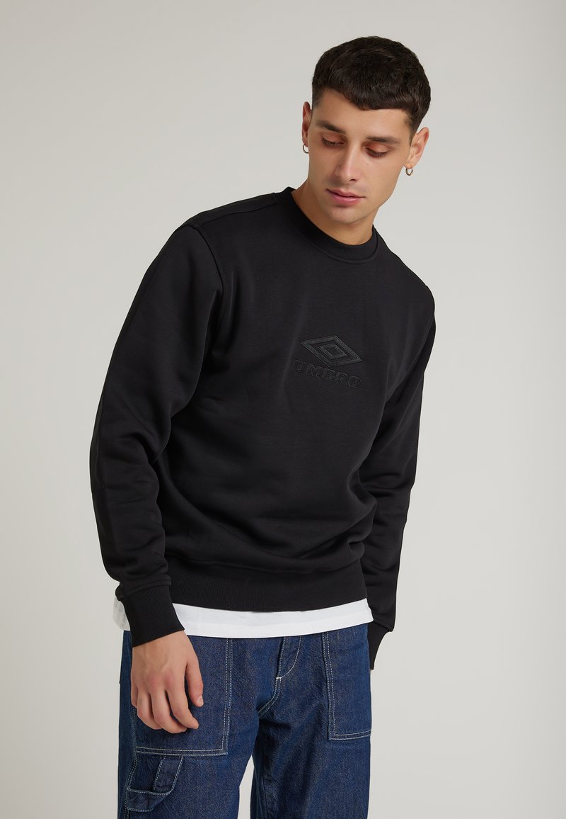 Giovane uomo che indossa un felpa nera Umbro e jeans blu, che guarda verso il basso a sinistra su uno sfondo chiaro e uniforme.