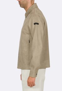 Giacca in suede beige con colletto, tasche laterali con zip e un accento nero sulla spalla sinistra, dotata di maniche lunghe e una vestibilità comoda.