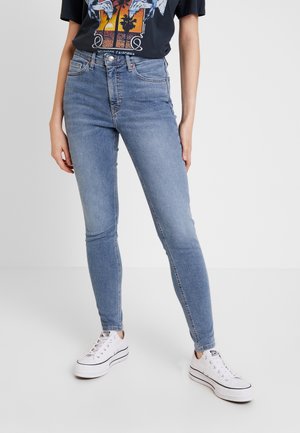 Personne portant un jean skinny bleu clair, un t-shirt graphique noir et des baskets basses blanches, debout sur un sol blanc.