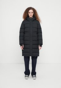 Μαύρο μπουφάν τύπου puffer με ψηλό γιακά, σχεδίαση με καπιτονέ και φερμουάρ μπροστά. Φορεμένο πάνω από μπλε σκούρο τζιν και λευκά αθλητικά παπούτσια. Λείο υλικό.