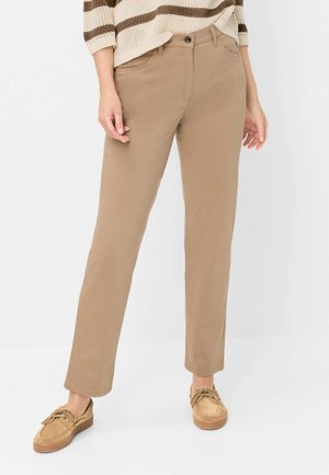 Kvinde iført beige bukser med lige ben, stribet beige og brun sweater samt lysebrune snørestøvler, stående mod en ensfarvet baggrund.