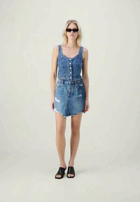 Γυναίκα φορώντας αμάνικο τζιν crop top με κουμπιά και κατεστραμμένη τζιν φούστα, μαύρες ανοιχτές μύτες μπαλαρίνες και σκούρα γυαλιά ηλίου, στέκεται σε απλό φόντο.