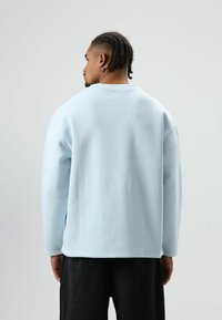 Sweat-shirt bleu clair à manches longues avec un col rond. Présente une texture lisse et une coupe décontractée, associé à un pantalon foncé.
