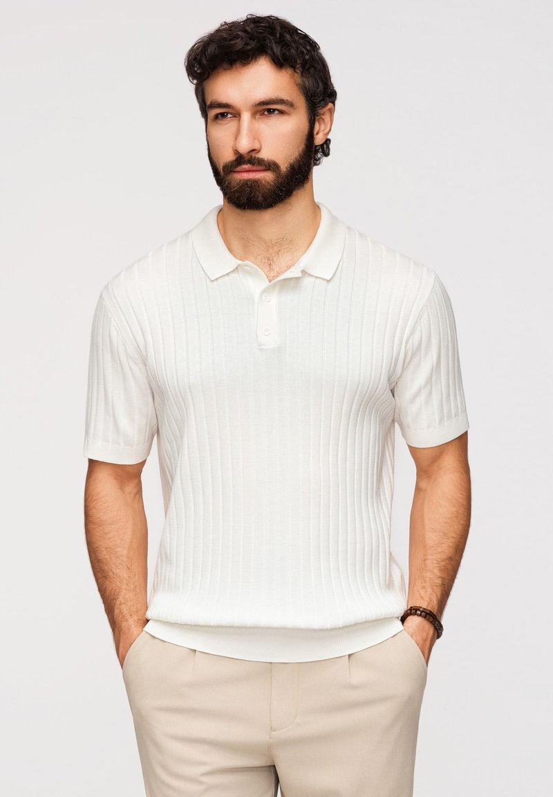 Homme barbu portant un polo blanc côtelé à manches courtes et un pantalon beige, debout les mains dans les poches sur fond blanc.