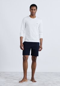 White long-sleeve shirt with black logo, paired with navy shorts. Model stands barefoot on a light carpeted surface. Neutral background.