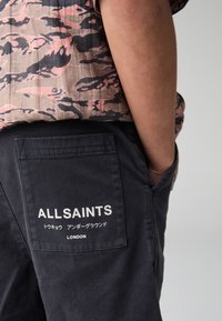 Čierne nohavice do pása z látky s náprsním vreckom s bielym logom "ALLSAINTS", doplnené o vzorovanú košeľu s krátkym rukávom v tlmených farbách.