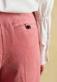 Rozā kordurojas bikses ar vertikāliem rievu rakstiem, aizmugurējā kabatā melns pogas aizdare, kopā ar baltu, savākta garām piedurknēm kreklu.