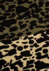 Tissu à motifs animaliers présentant un mélange de taches noires et vert olive sur un fond beige. La texture semble douce et duveteuse.