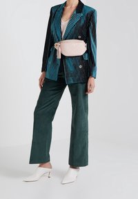 Blazer rayé noir et teal sur un haut blanc, pantalon en velours côtelé vert et mules blanches. Sac banane matelassé rose avec un accent à frange.