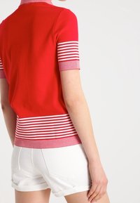 Červený top s krátkým rukávem, bílými vodorovnými pruhy na spodním lemu a manžetách, s celým červeným tělem. V kombinaci s bílými šortkami.