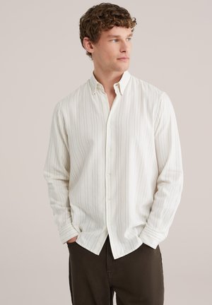 Homme aux cheveux bouclés portant une chemise blanche à manches longues avec de fines rayures verticales, mains dans les poches, regardant sur le côté.