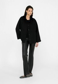 Zwarte oversized jas met een getextureerde kraag, grote knopen aan de voorkant, gecombineerd met strakke zwarte neplederen broek en grijze instapschoenen.