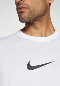 Camiseta de algodón blanca con un cuello redondo y mangas cortas, que presenta un prominente logo de Nike en negro en el pecho.