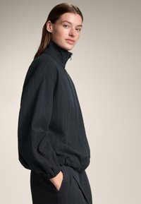 Fekete, bordázott cipzáras dzseki magas gallérral, laza szabással és rugalmas mandzsettákkal. Függőleges vékony csíkok enyhén kontrasztos színben.