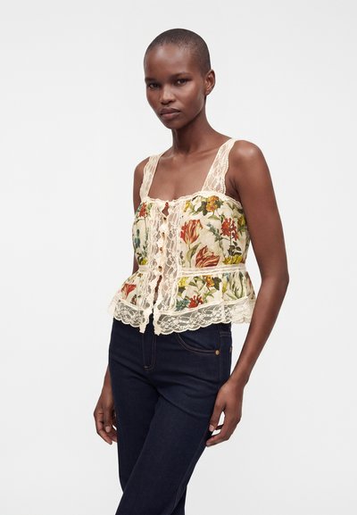 Femme portant un haut fleuri sans manches avec un bord en dentelle et un jean foncé, debout devant un fond clair uni.