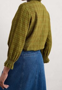 Blouse à carreaux verts avec poignets smockés, associée à une jupe en denim bleu. La blouse a une coupe décontractée et une texture légère.