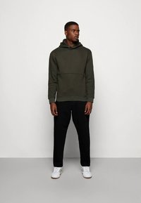 Uomo con un hoodie verde scuro, pantaloni neri e sneakers bianche in piedi contro un muro bianco semplice, che guarda di lato.